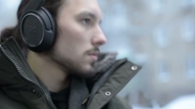 Bir ceket portre genç uzun saçlı sakallı adam kapatın ve büyük kulaklıklar pencerenin tramvayla toplu taşıma oturur ve müzik veya bir sesli kitap için kışın dinler