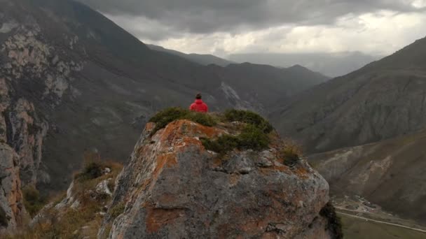 Un joven se sienta en una posición de loto en la cima de una roca en las montañas y medita. Vista aérea. vuelo a. vista desde atrás — Vídeo de stock