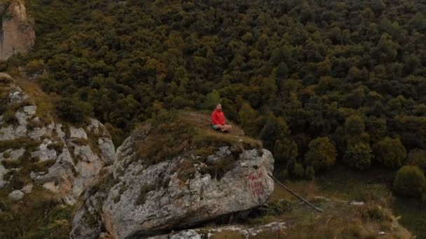 Um jovem senta-se em uma posição de lótus no topo de uma rocha nas montanhas e medita. Vista aérea. voo ao redor — Vídeo de Stock