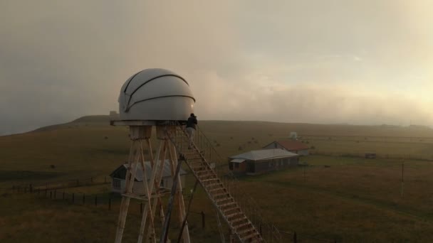 一个旅行者摄影师女孩站在高加索天文台的自主望远镜的梯子上, 手里拿着相机, 拍摄日落的照片。鸟瞰 — 图库视频影像
