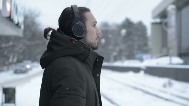 Portrait d'un jeune homme aux cheveux longs avec une barbe dans un casque debout à un arrêt de tram en hiver et attendant un tram écoutant de la musique — Video