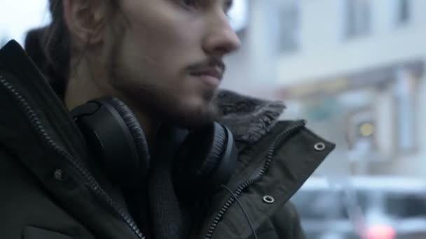 穿着夹克和大耳机的年轻长头发的胡须男子坐在靠窗的电车旁的公共交通中, 在冬天听音乐或有声读物 — 图库视频影像