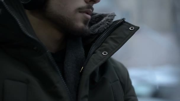 Retrato de cerca Joven hombre barbudo de pelo largo con chaqueta y auriculares grandes se sienta en el transporte público en el tranvía por la ventana y escucha música o un audiolibro en el invierno — Vídeos de Stock