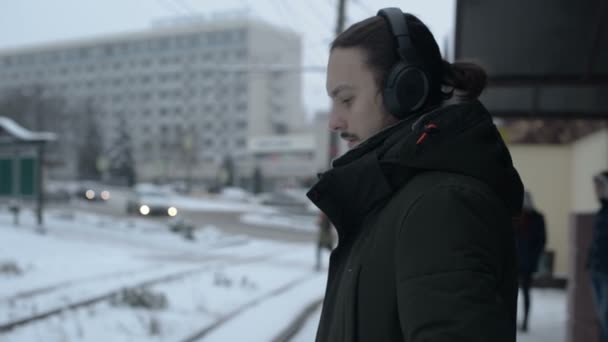 Портрет молодого длинноволосого человека с бородой в наушниках, стоящего на остановке трамвая зимой и ожидающего трамвая, слушающего музыку — стоковое видео