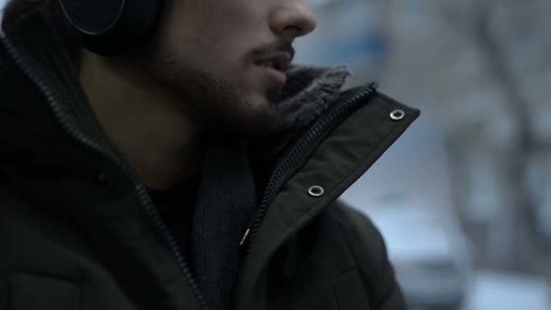 Close-up van portret jonge langharige bebaarde man in een jasje en grote hoofdtelefoon zit in openbaar vervoer door de tram bij het raam en luistert naar muziek of een audioboek in de winter — Stockvideo