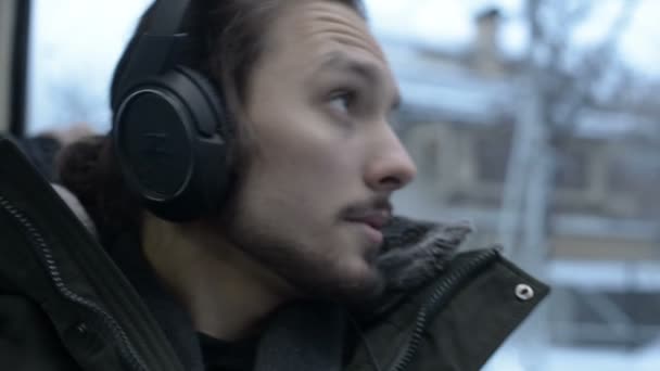 Retrato de cerca Joven hombre barbudo de pelo largo con chaqueta y auriculares grandes se sienta en el transporte público en el tranvía por la ventana y escucha música o un audiolibro en el invierno — Vídeo de stock