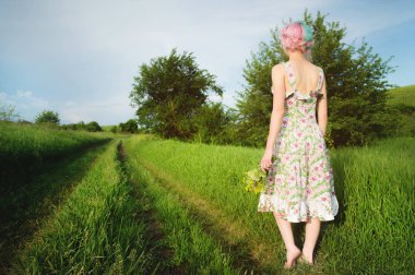 Hafif pamuklu elbise sevimli neşeli genç kız gün batımında yaz yeşil zemin karşı ülke yol boyunca elini kolunu sallaya dönüyor. Arkadan görüntülemek