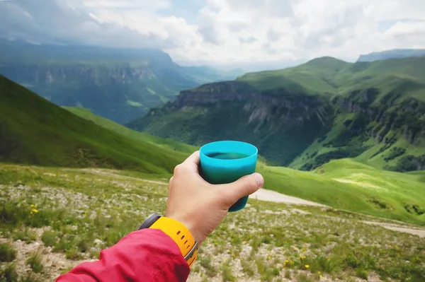First-person view of a mans hand holding a plastic cup of tea against a plateau of green hills and a cloudy sky in summer — Stock Photo, Image