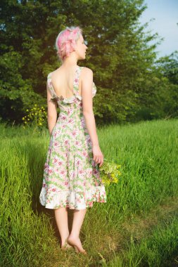 Hafif pamuklu elbise sevimli neşeli genç kız gün batımında yaz yeşil zemin karşı ülke yol boyunca elini kolunu sallaya dönüyor. Arkadan görüntülemek