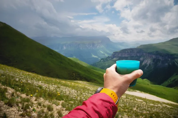 First-person view of a mans hand holding a plastic cup of tea against a plateau of green hills and a cloudy sky in summer — Stock Photo, Image