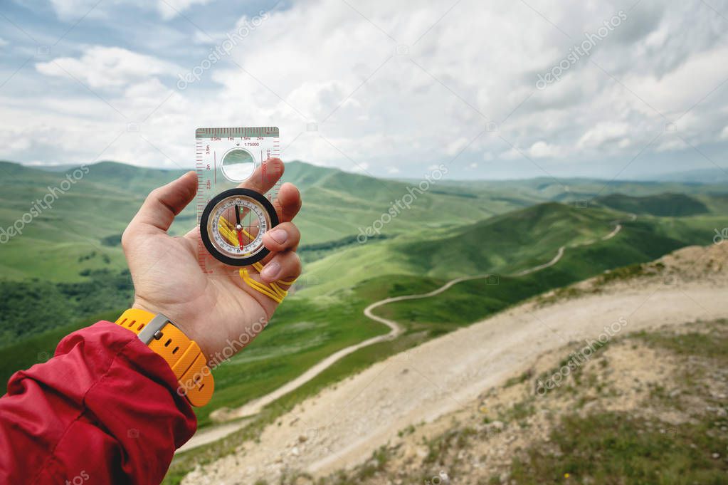 Male hand is holding a magnetic compass on the background of hills and the sky with clouds. The concept of traveling and finding your life path