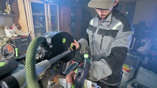 Um jovem com barba em macacão cinza por profissão um carpinteiro trabalha com uma máquina de corte circular em sua oficina em casa. Corte de madeira — Vídeo de Stock
