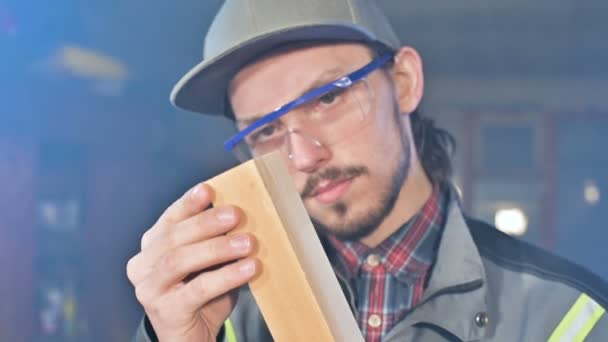 Geconcentreerde jonge bebaarde meubelmaker in bril en een cap controleert het werkstuk voor geschiktheid tijdens het bedrijf dat in zijn handen en onderzoeken — Stockvideo