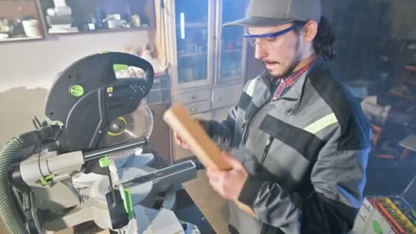 Joven carpintero barbudo concentrado en gafas y una gorra comprueba la idoneidad de la pieza de trabajo mientras la sostiene en sus manos y examina — Vídeos de Stock