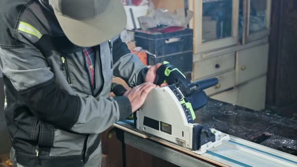 Молодой человек с бородой в сером комбинезоне по профессии плотник работает с круглой резкой машиной в своей домашней мастерской. Резка древесины — стоковое видео