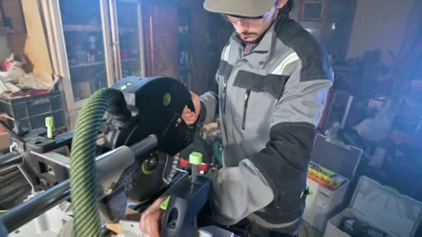 Een jonge man met een baard in grijze Amerikaanse overalls van beroep timmerman werkt met een cirkelvormige snijmachine in zijn huis atelier. Hout snijden — Stockvideo