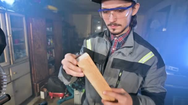 Concentrated young bearded joiner in goggles and a cap checks the workpiece for suitability while holding it in his hands and examining — Stock Video