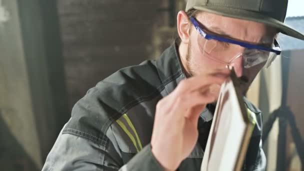 Joven carpintero barbudo concentrado en gafas y una gorra comprueba la idoneidad de la pieza de trabajo mientras la sostiene en sus manos y examina — Vídeo de stock