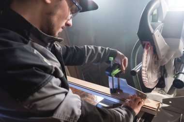 Bir kap mesleği marangoz gri bir ceket giyen genç esmer adam ahşap kurulları daire testere ile bir atölye tezgah tablosunda keser. Profesyonel ekipman ev atölye çalışmaları