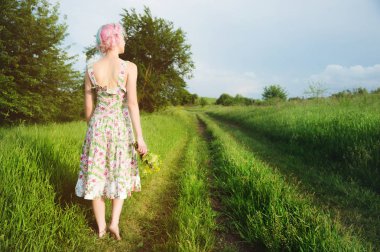 Hafif pamuklu elbise sevimli neşeli genç kız gün batımında yaz yeşil zemin karşı ülke yol boyunca elini kolunu sallaya dönüyor. Arkadan görüntülemek