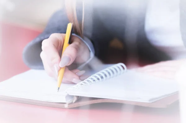 Close-up of a female students hand in a cafe behind the glass with a reflection that writes something with a ballpoint pen in a notebook. Decontrast for reflection