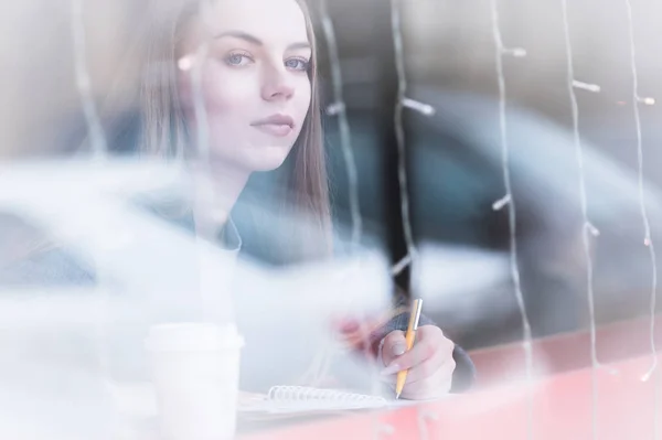 Portrét dívky atraktivní mladý novinář s pero a Poznámkový blok v kavárně za vitríny. Non kontrastní zobrazení prostřednictvím okna odraz ve skle. Při pohledu na fotoaparát — Stock fotografie