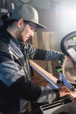 Bir kap mesleği marangoz gri bir ceket giyen genç esmer adam ahşap kurulları daire testere ile bir atölye tezgah tablosunda keser. Profesyonel ekipman ev atölye çalışmaları