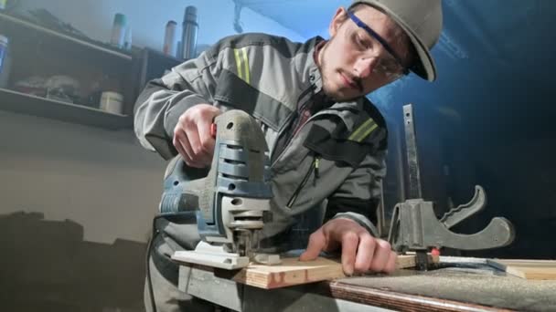 Young man with a beard in gray overalls and goggles by profession a carpenter works as an electric jigsaw in his home workshop. Cutting wooden parts — Stock Video