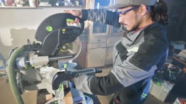 Mesleği gri Tulum içinde sakallı genç bir adam onun ev atölye bir dairesel kesme makinesi ile bir marangoz çalışır. Ahşap kesim