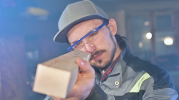 Jeune menuisier barbu concentré dans des lunettes et un capuchon vérifie l'aptitude de la pièce tout en la tenant dans ses mains et en examinant — Video