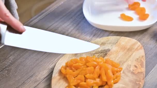 Крупный план женских рук, режущих сушеные абрикосы ножом на разделочной доске. Вегетарианская кухня — стоковое видео