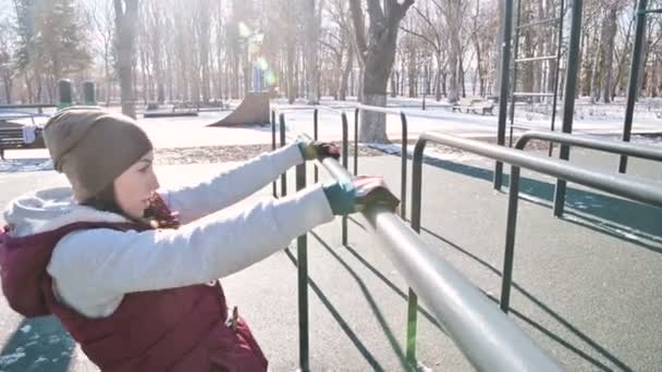 Portrait d'une athlète fille portant un chapeau avec des gants et un gilet chaud faisant des exercices sur des bars inégaux sur un terrain de sport en plein air en hiver par une journée ensoleillée — Video