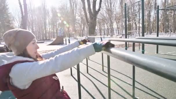 Retrato de una atleta en un sombrero con guantes y un chaleco caliente haciendo ejercicios en bares irregulares en un campo de deportes al aire libre en invierno en un día soleado — Vídeos de Stock