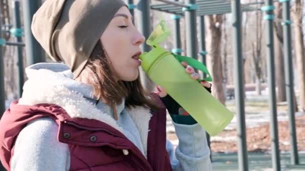 Sport flicka på en utomhus träning i en park på idrottsplanen i vintern på en solig dag dricksvatten från en shaker — Stockvideo