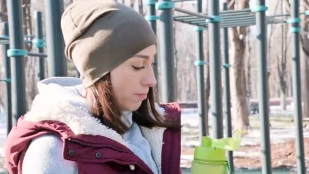 Sports girl at an outdoor workout in a park on the sports field in the winter on a sunny day drinking water from a shaker — Stock Video