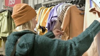 Bir kız bir yeşil kazak ve sarı şapka şeyler mağazası aracılığıyla yürüyor ve ne satın almak için seçer. Askıları şeylerde dokunur ve fiyat etiketleri görünüyor