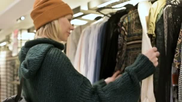 Una chica con un suéter verde y un sombrero amarillo camina a través de una tienda de cosas y elige qué comprar. Toca cosas en perchas y mira etiquetas de precios — Vídeos de Stock