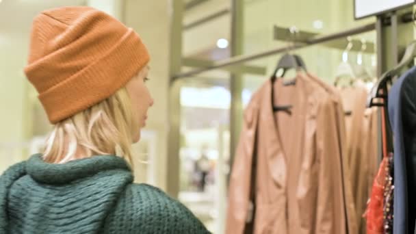 Una chica con un suéter verde y un sombrero amarillo camina a través de una tienda de cosas y elige qué comprar. Toca cosas en perchas y mira etiquetas de precios — Vídeo de stock