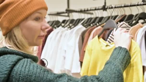 Bir kız bir yeşil kazak ve sarı şapka şeyler mağazası aracılığıyla yürüyor ve ne satın almak için seçer. Askıları şeylerde dokunur ve fiyat etiketleri görünüyor — Stok video