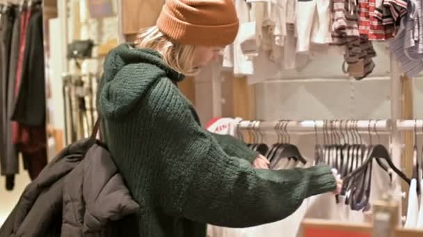 Девушка в зеленом свитере и в жёлтой шляпе проходит по магазину вещей и выбирает, что купить. Касается вещей на вешалки и смотрит на ценники — стоковое видео