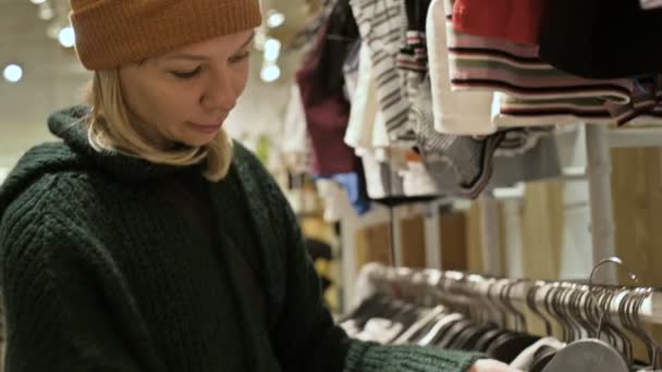 Una chica con un suéter verde y un sombrero amarillo camina a través de una tienda de cosas y elige qué comprar. Toca cosas en perchas y mira etiquetas de precios — Vídeos de Stock