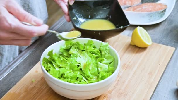 Pendekatan tangan perempuan di dapur rumah ditambahkan pada saus salad daun vegetarian dan dicampur dengan sendok pada papan potong kayu — Stok Video