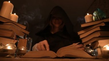 Mum ışığı ve seyir için bir kitap üzerinde bir büyü tarafından karanlık bir odada bir başlıklı orta yakın çekim kız sihirbaz. Düşük anahtar. Mistik küçük Dof