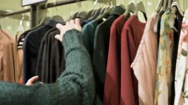 Bir kız bir yeşil kazak ve sarı şapka şeyler mağazası aracılığıyla yürüyor ve ne satın almak için seçer. Askıları şeylerde dokunur ve fiyat etiketleri görünüyor