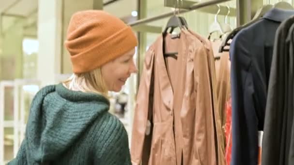 Een meisje in een groene trui en een gele hoed begeleidt bij een winkel van dingen en kiest wat te kopen. Raakt dingen op hangers en kijkt naar de prijskaartjes — Stockvideo