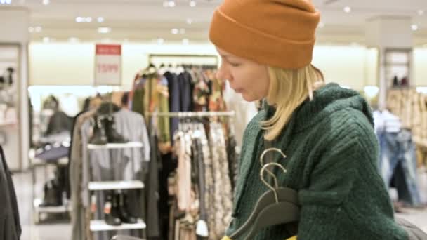 Een meisje in een groene trui en een gele hoed begeleidt bij een winkel van dingen en kiest wat te kopen. Raakt dingen op hangers en kijkt naar de prijskaartjes — Stockvideo