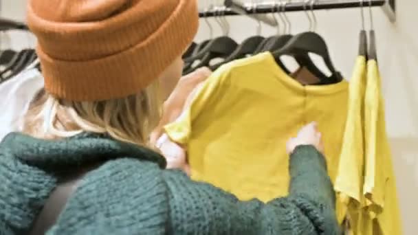 Dziewczyna w zielony sweter i żółty kapelusz idzie w sklepie rzeczy i wybiera co kupić. Dotyka rzeczy na wieszakach i patrzy na metki — Wideo stockowe