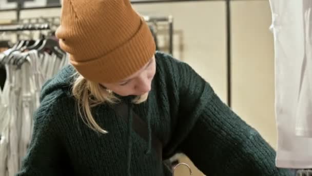 Uma menina com uma camisola verde e um chapéu amarelo caminha por uma loja de coisas e escolhe o que comprar. Toca coisas em cabides e olha para etiquetas de preço — Vídeo de Stock