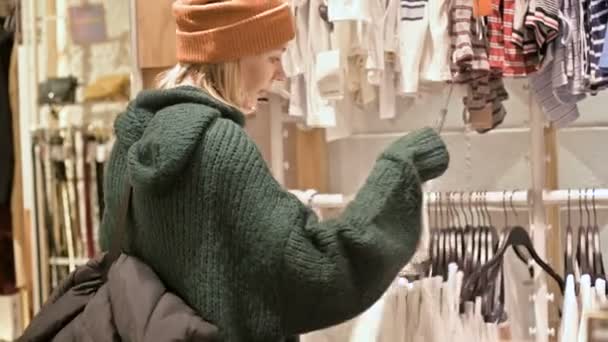 Uma menina com uma camisola verde e um chapéu amarelo caminha por uma loja de coisas e escolhe o que comprar. Toca coisas em cabides e olha para etiquetas de preço — Vídeo de Stock