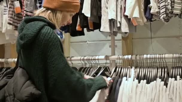 Девушка в зеленом свитере и в жёлтой шляпе проходит по магазину вещей и выбирает, что купить. Касается вещей на вешалки и смотрит на ценники — стоковое видео
